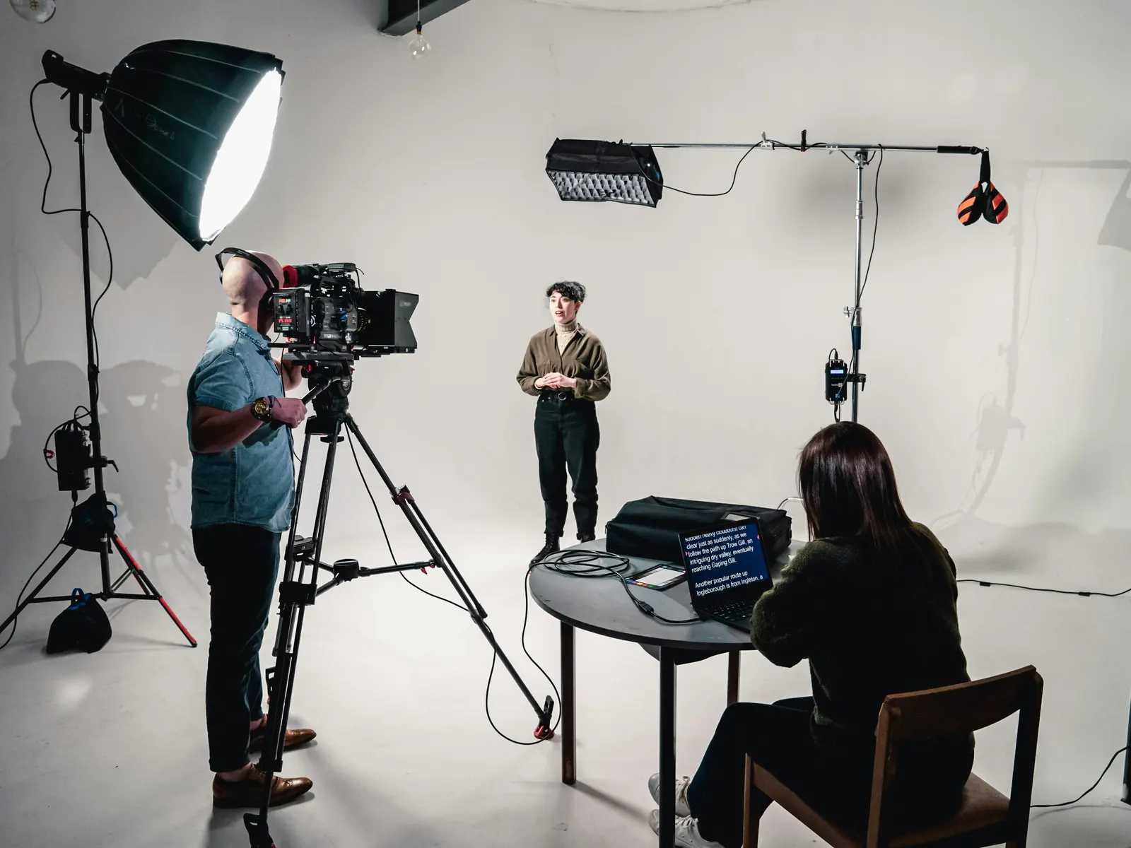Camera operator filming a presenter in a bright TV studio while a producer runs the teleprompter