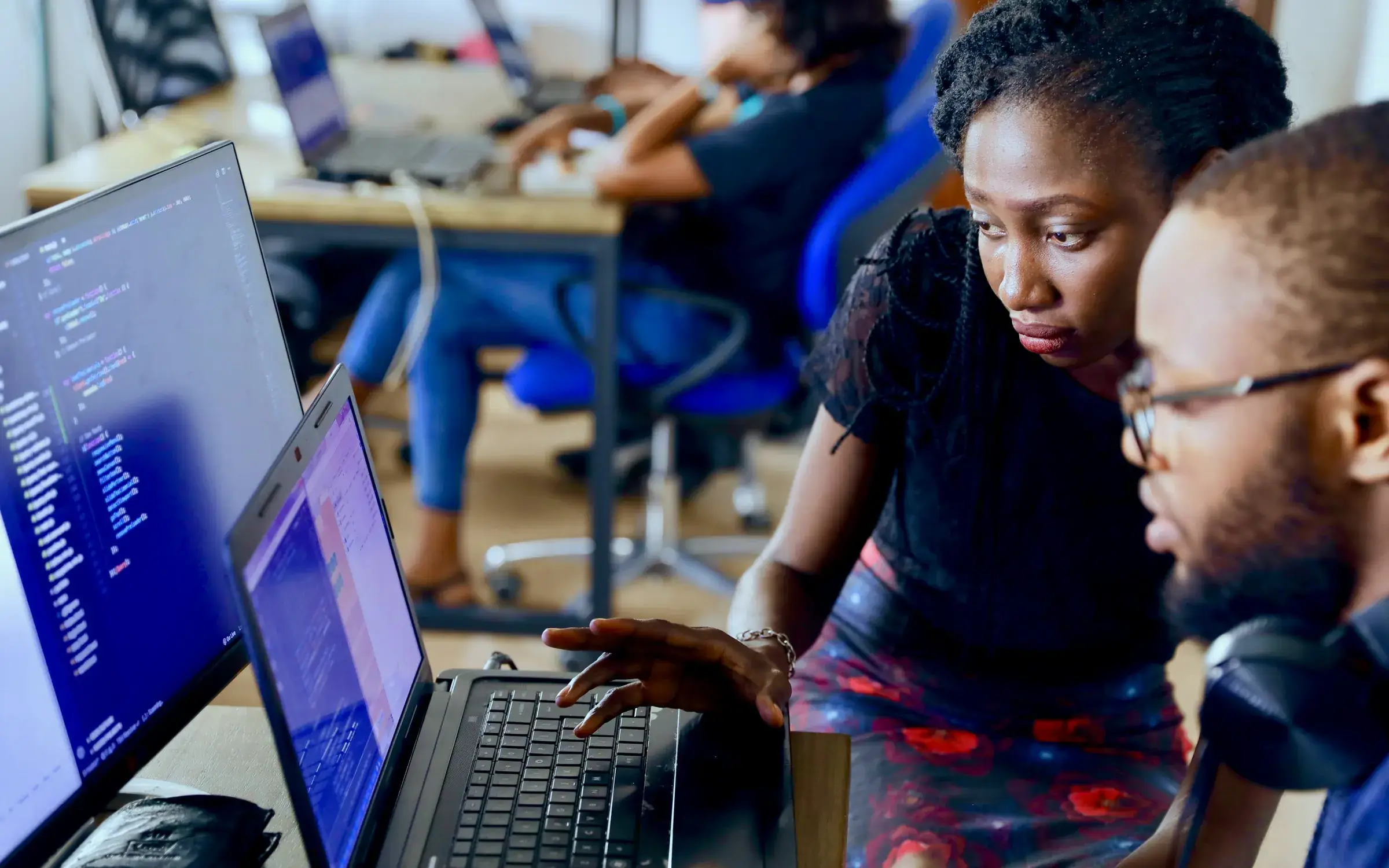 Two engineers reviewing code together on a laptop in a busy team workspace